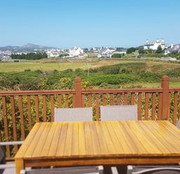 An outdoor wooden table with chairs on a balcony overlooking fields and houses at Windy Ridge in Trearddur Bay