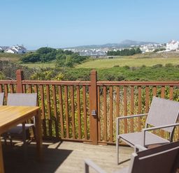 An outdoor wooden deck with table and chairs overlooking green fields and houses in the distance at Windy Ridge in Trearddur Bay