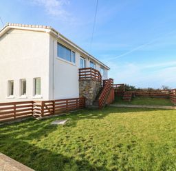 A fenced yard with grass and wooden stairs leading to a white two-story house at Windy Ridge in Trearddur Bay