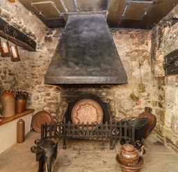 An old stone room with a large fireplace a black iron fireguard copper utensils and pottery jars at Field View in Curland near Taunton