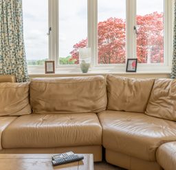 A beige leather sectional sofa in front of a window with blue patterned curtains and a wooden table with remote controls at Hazelseat House in Graythwaite near Ulverston