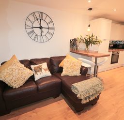 A living room with a brown leather sofa and cushions next to a kitchen with white cabinets at Swale Cottage in Beadnell