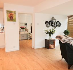A room with wooden flooring a black armchair with a blanket an american flag patterned side table with flowers a wall art a door with wooden blinds and a kitchen area with white cabinets at Swale Cottage in Beadnell