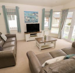 A living room with two brown sofas a coffee table a TV on a stand and windows with curtains at Cwtch Lodge 42 Stepaside near Wisemans Bridge