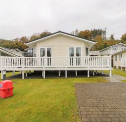 A beige lodge with white railings and multiple windows on a grassy plot next to a paved driveway at Cwtch Lodge 42 Stepaside near Wisemans Bridge