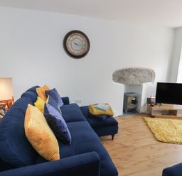 A living room with a blue sofa and yellow cushions a clock on the wall a television on a wooden stand and a yellow rug at Bryn Derfel in Borth-y-Gest near Porthmadog
