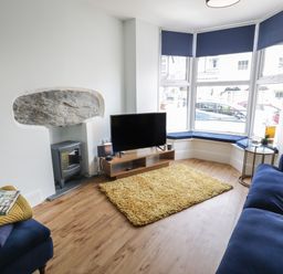A living room with blue sofas a yellow rug a television on a wooden stand and a bay window with blue blinds at Bryn Derfel in Borth-y-Gest near Porthmadog