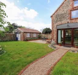 A garden with a gravel path leading to a stone building with glass doors and an outdoor seating area at The Little Hay Barn in Bacton