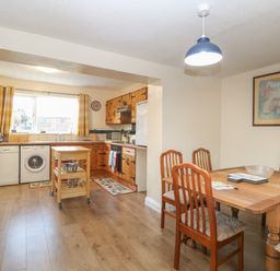 A kitchen with wooden cabinets, a small kitchen island, dining table with chairs, and a window with yellow curtains at Rose Cottage in Downham Market