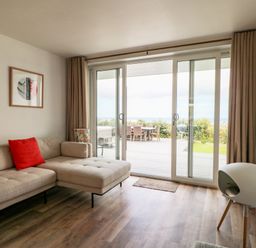 A living room with a beige sectional sofa with red cushions and a sliding glass door opening to a patio at Pepperpots in Mawgan Porth