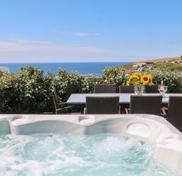 An outdoor seating area with a hot tub a dining table with chairs sunflowers and a bottle of wine with glasses overlooking the sea at Pepperpots in Mawgan Porth