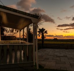 A porch with wooden railing overlooking a paved area and grassy field with a palm tree at sunset at Bennar Fawr in Dyffryn Ardudwy