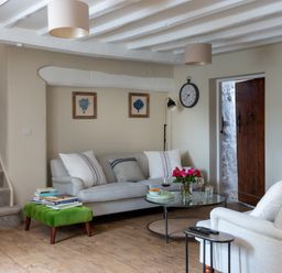 A living room with sofas a green ottoman with books a glass coffee table flowers and a clock on the wall at Bennar Fawr in Dyffryn Ardudwy