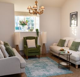 A living room with two beige sofas a green armchair two lamps a chandelier and a window with flowers at Bennar Fawr in Dyffryn Ardudwy