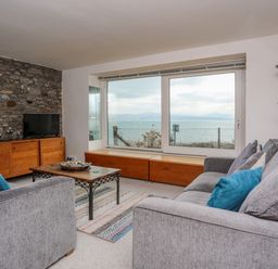 A living room with two gray sofas a wooden cabinet a flat screen tv and a large window with sea view at Awel Y Mor in Criccieth