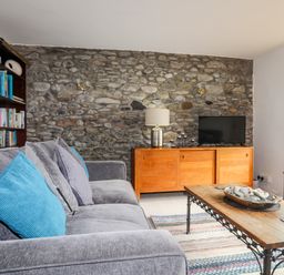 A living room with a grey sofa with blue and grey cushions a wooden coffee table with stones a wooden cabinet with a lamp and a TV a stone wall and a bookshelf at Awel Y Mor in Criccieth