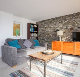A living room with a gray sofa and colorful cushions a wooden coffee table a wooden cabinet with a lamp and television a bookshelf and a stone wall at Awel Y Mor in Criccieth