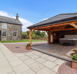 An outdoor area with a stone house, a wooden gazebo, a hot tub, a bench, potted plants, and a paved walkway at Plas Iolyn in Pentrefoelas near Betws-Y-Coed