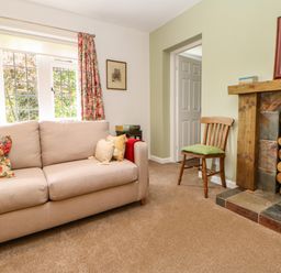A living room with a beige sofa and floral cushions a wooden chair with a green cushion a fireplace with stacked logs and tiled hearth at The Coach House Shepherd's Dene near Corbridge