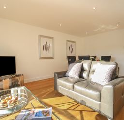 A living room with a leather sofa pillows a glass coffee table with mugs and pastries a flat screen tv and dining chairs at Harbourside Haven Apartment 2 in Weymouth