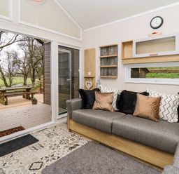A living room with a sofa with cushions a wall clock and shelving and a sliding glass door opening to an outdoor picnic table at Woodland Escape in Rhayader