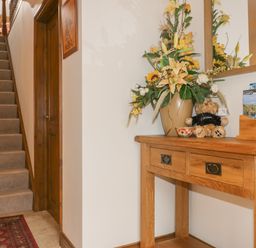 A hallway with a carpeted staircase wooden door a wooden table with a flower arrangement a teddy bear and brochures at Meadowside in Goonhavern