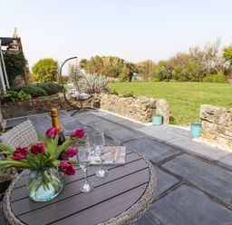 A patio with a round table holding flowers wine glasses and a bottle a hanging chair and a stone wall with grass beyond at The Roost in Porthallow