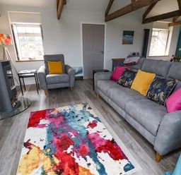 A living room with a grey sofa and armchair with colorful cushions a multicolored rug and a wood burning stove at The Roost in Porthallow