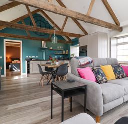 An open living space with exposed wooden beams a grey couch with colorful cushions a dining table with black chairs and a kitchen area with teal walls at The Roost in Porthallow
