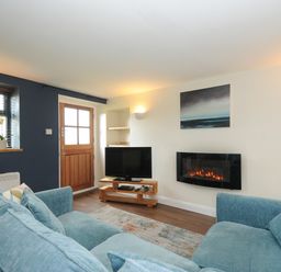 A living room with blue sofas a wooden door a television and a wall mounted electric fireplace at The Bower in Manaccan near St Keverne
