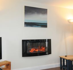 A room with a wall-mounted electric fireplace a television on a wooden stand a painting above the fireplace a wooden floor lamp and a small wooden table at The Bower in Manaccan near St Keverne