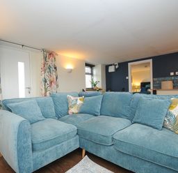 A living room with a blue sectional sofa cushions floral curtains and a kitchen area at The Bower in Manaccan near St Keverne