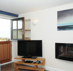 A corner of a living room with a wooden door open above blue wall section a flat screen tv on a wooden stand and an electric fireplace on a white wall at The Bower in Manaccan near St Keverne