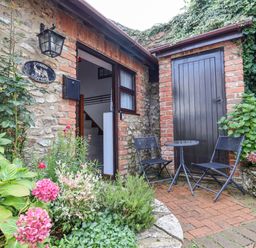 An outdoor seating area with two black chairs and a small round table next to a wooden door with plants and flowers outside at Coach Cottage in Colyton