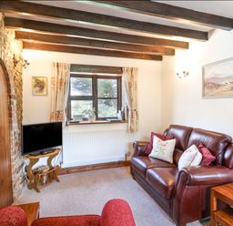 A living room with a leather sofa a wooden TV stand a stone wall with an arched wooden door and exposed ceiling beams at Coach Cottage in Colyton