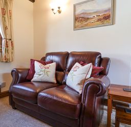 A leather sofa with floral and plain cushions next to a wooden side table under a wall painting at Coach Cottage in Colyton