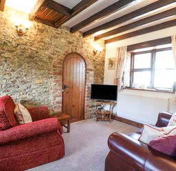 A living room with a stone wall wooden door red fabric armchair brown leather sofa and a TV on a wooden stand at Coach Cottage in Colyton