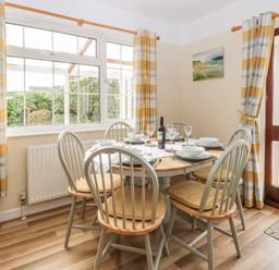A dining area with a round wooden table set for six and chairs near windows and a door at Doves Nest in Martham