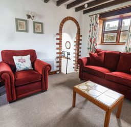 A living room with red armchair and sofa a wooden coffee table and floral curtains at Stable Cottage in Colyton