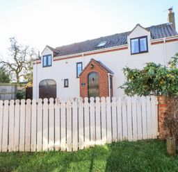 A white house with brown windows and doors behind a white wooden fence with grass in front at Anvil Cottage in Appleton Wiske