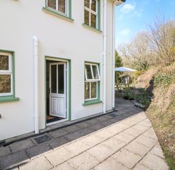 An exterior view of a white house with green window frames and an open door next to a paved pathway leading to a garden area with a picnic table and umbrella at 1 Closheen Lane in Rosscarbery County Cork