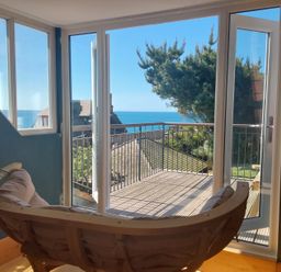 A room with a curved wooden chair facing glass doors leading to a balcony with a sea view at Sandy Combe in West Bay