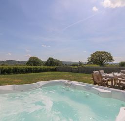 An outdoor hot tub with bubbling water and wooden dining table with chairs on a patio in a grassy yard at Hill Radnor - The Sheepfold in Llanyre near Llandrindod Wells