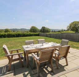 A wooden outdoor dining table with six chairs on a stone patio in a garden with trees and hedges at Hill Radnor - The Sheepfold in Llanyre near Llandrindod Wells
