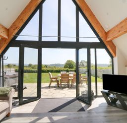 A room with a sofa a television and large windows overlooking a patio and garden at Brecknock Cheviot - The Sheepfold in Llanyre near Llandrindod Wells