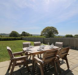 A wooden outdoor dining table with six chairs set with dishes and glasses on a stone patio overlooking a fenced grassy yard at Brecknock Cheviot - The Sheepfold in Llanyre near Llandrindod Wells