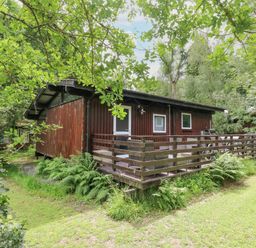 A wooden cabin with a fenced porch surrounded by trees and grass at Doirewood Penlan Holiday Park near Cenarth