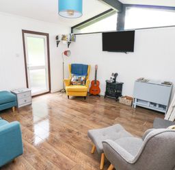 A living room with a blue sofa a yellow armchair a grey armchair with footstool a wall mounted television and a guitar at Doirewood in Penlan Holiday Park near Cenarth