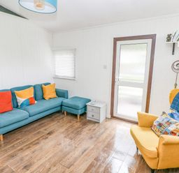 A living room with a blue sofa with colorful cushions a yellow armchair with cushions and a blue throw a small side table and a wooden floor at Doirewood Penlan Holiday Park near Cenarth