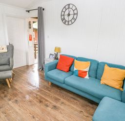 A living room with a blue sofa with cushions a grey armchair with footstool a side table with a lamp and wall clock at Doirewood Penlan Holiday Park near Cenarth